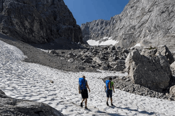 zwei Personen queren ein Schneefeld umgeben von felsigem Gelände am Berg